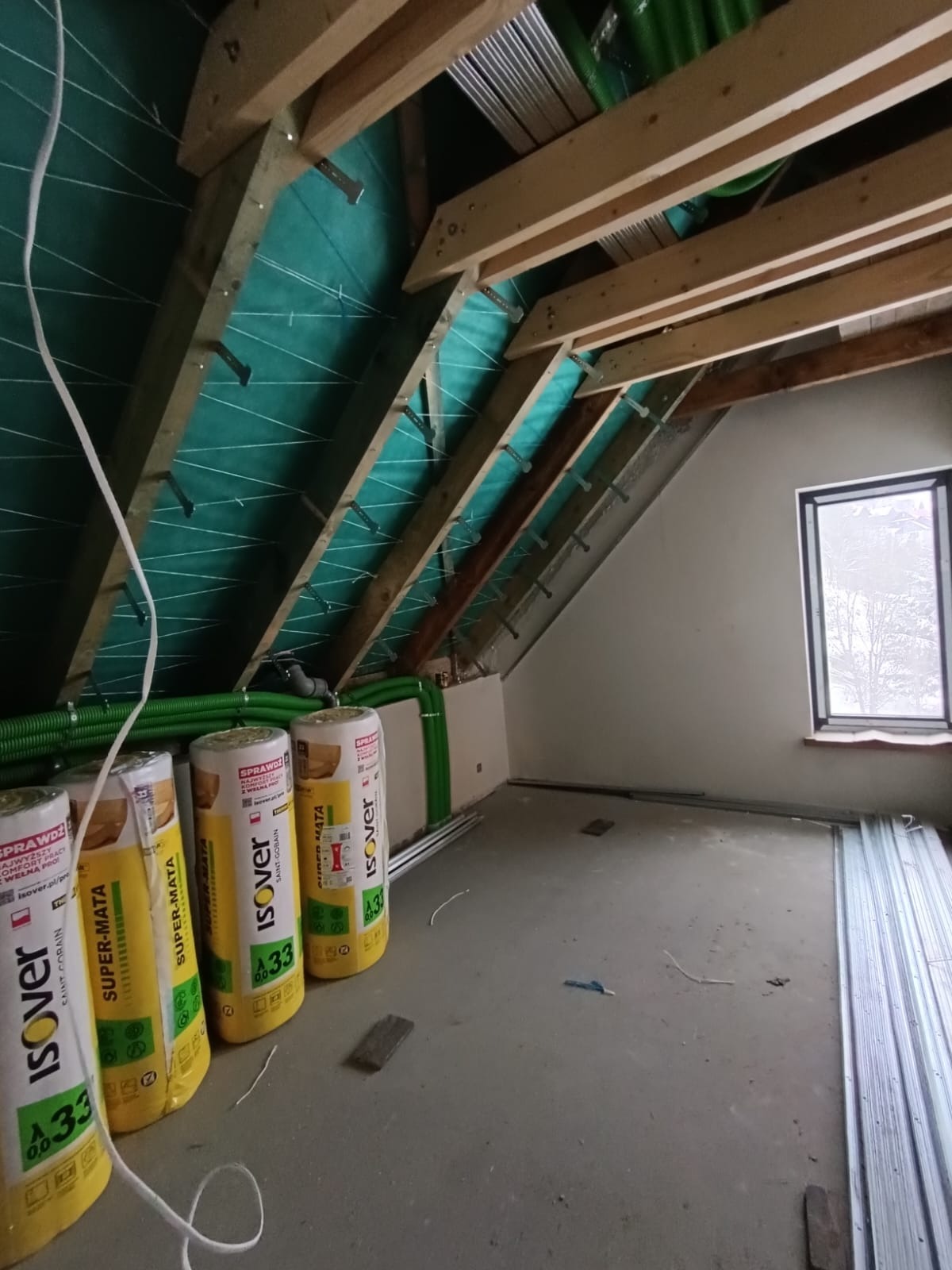 Poddasze w trakcie ocieplania: widoczne rolki wełny mineralnej Isover Super-Mata A33, zielone rury wentylacyjne, drewniana konstrukcja dachu z folią paroizolacyjną i metalowe profile.