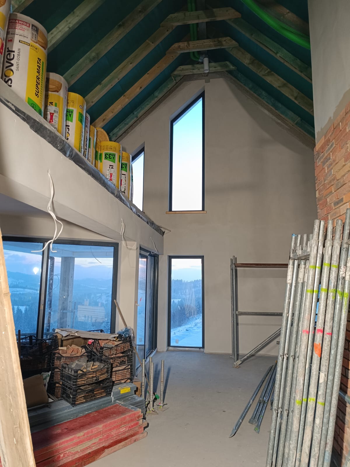 Wnętrze domu w stanie surowym zamkniętym z widokiem na zaśnieżony krajobraz przez duże okna; widoczne ocieplenie w rolkach na górnej kondygnacji, rusztowania i fragment murowanej ściany z cegły.