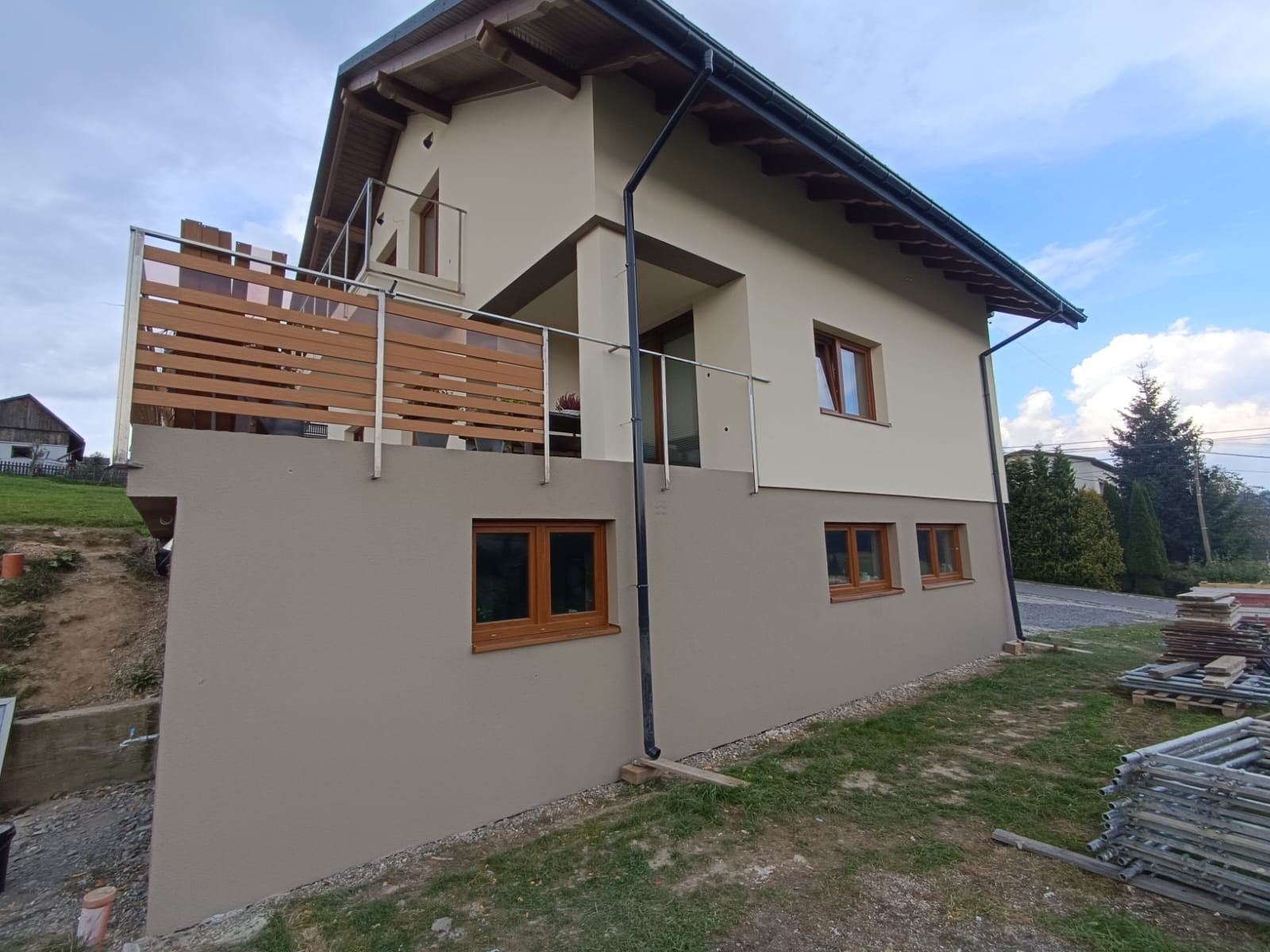 Dwukondygnacyjny dom z beżową elewacją i drewnianymi oknami, balkon z drewnianą balustradą, widoczne rusztowania i teren w trakcie zagospodarowania.