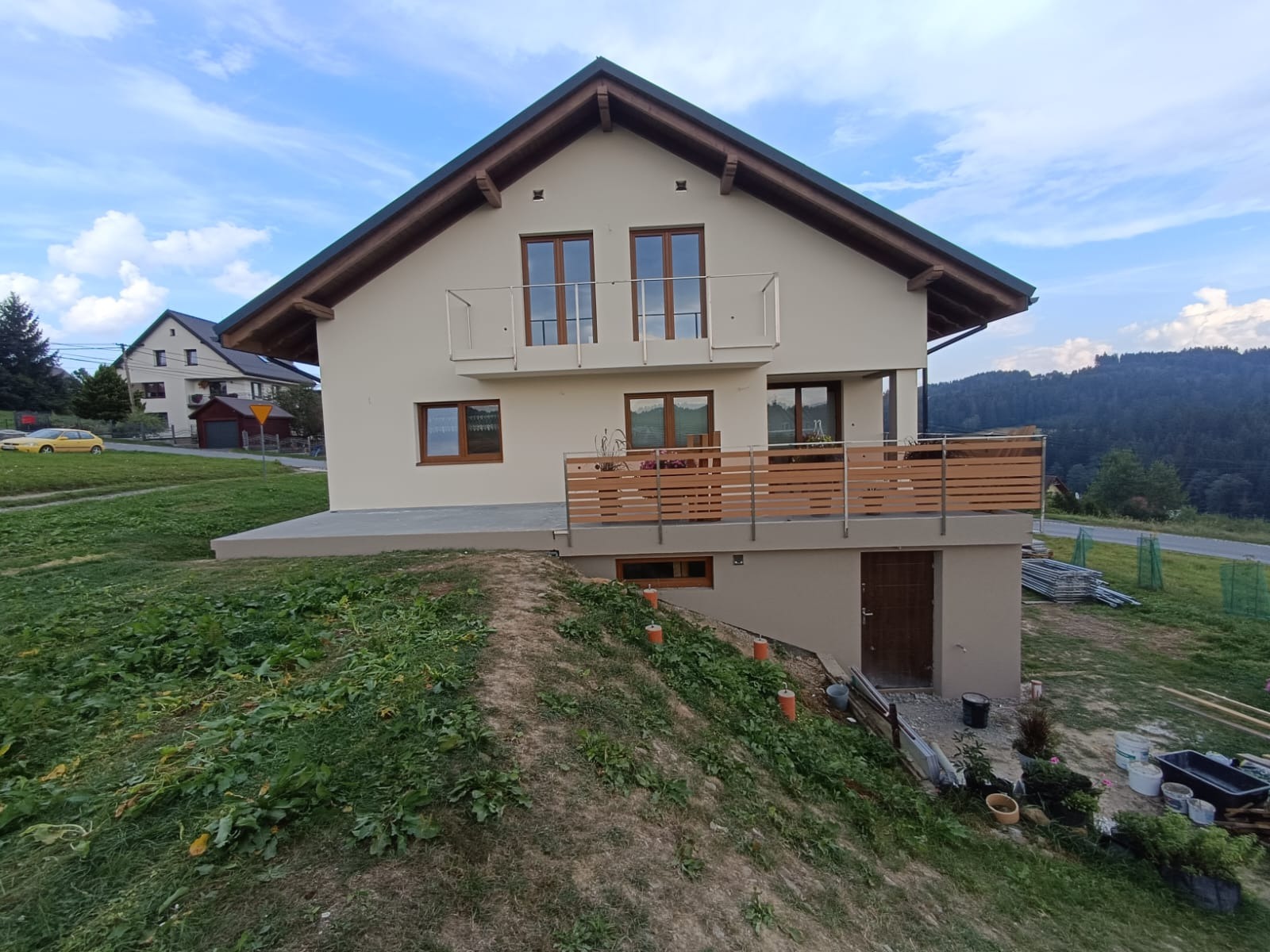 Nowoczesny, dwukondygnacyjny dom z beżową elewacją, balkonem z metalową balustradą i drewnianym wykończeniem, widoczny z perspektywy zbocza z rozkopaną ziemią i materiałami budowlanymi...