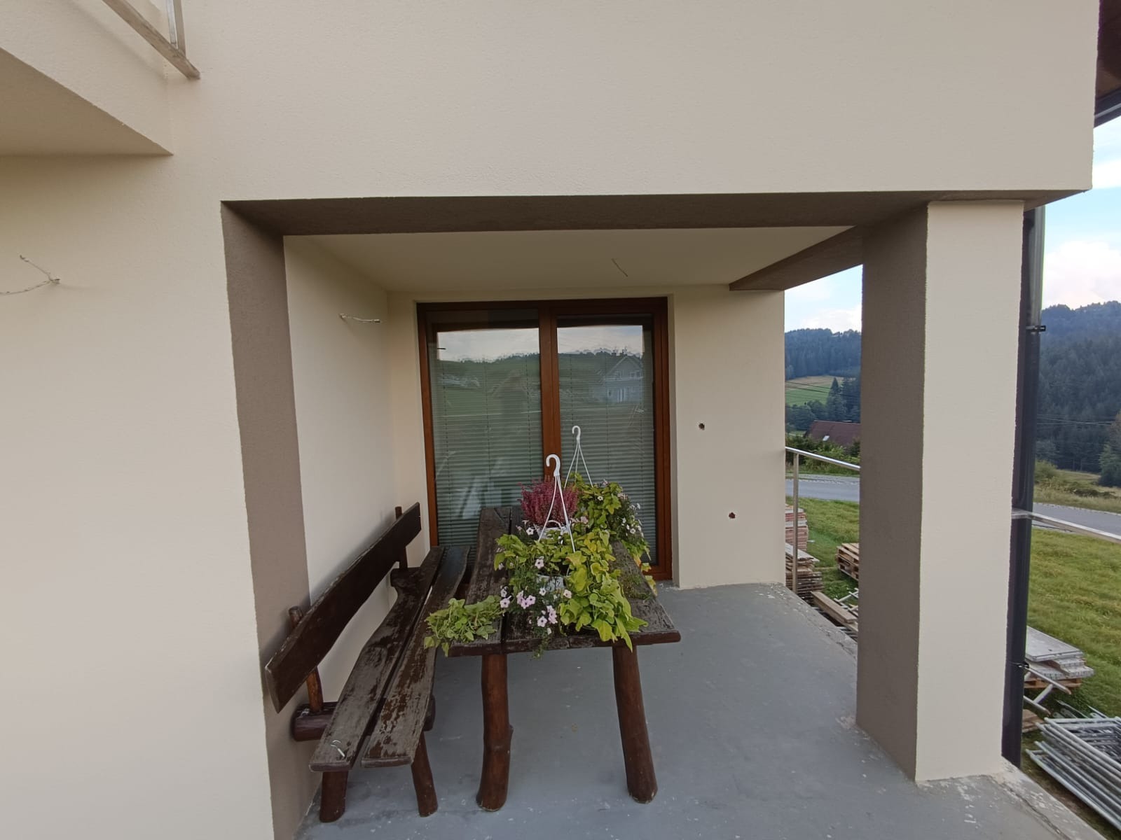 Zadaszony taras domu jednorodzinnego z drewnianym stołem i ławką porośniętymi roślinami, widok na górzysty krajobraz w tle.