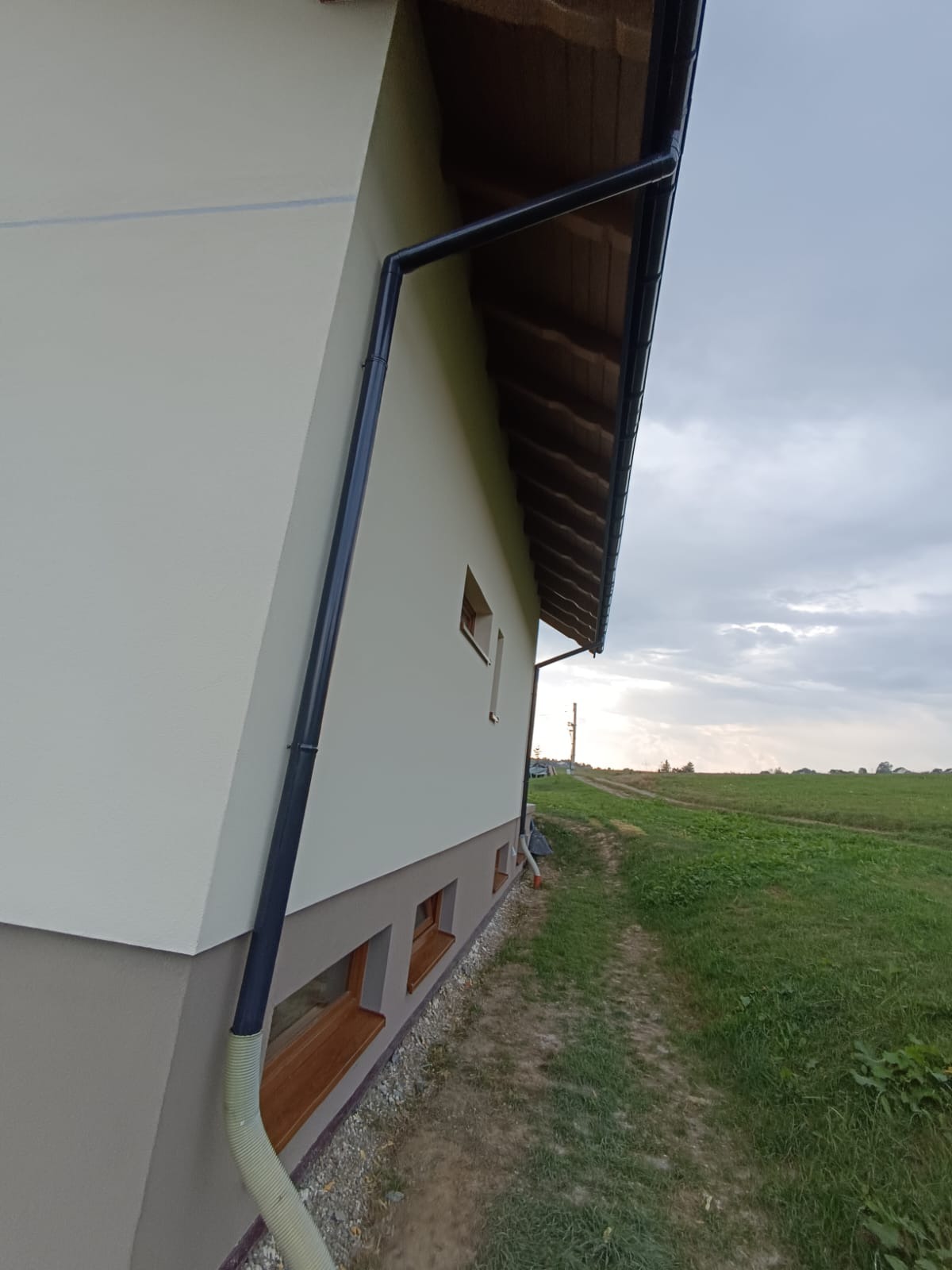 Świeżo otynkowana ściana domu z zamontowaną rynną spustową i widocznym fragmentem izolacji fundamentów, na tle zielonego pola i pochmurnego nieba.