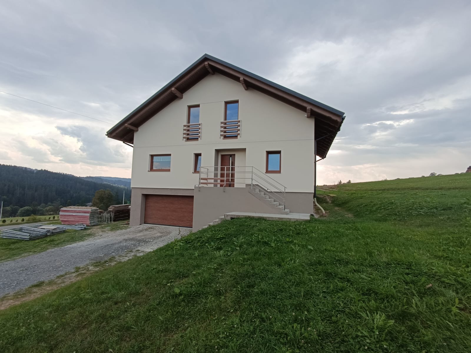 Dwupiętrowy dom z garażem wbudowanym w zbocze, z drewnianą stolarką okienną i schodami z metalową barierką, na tle zielonego wzgórza i zalesionych gór.