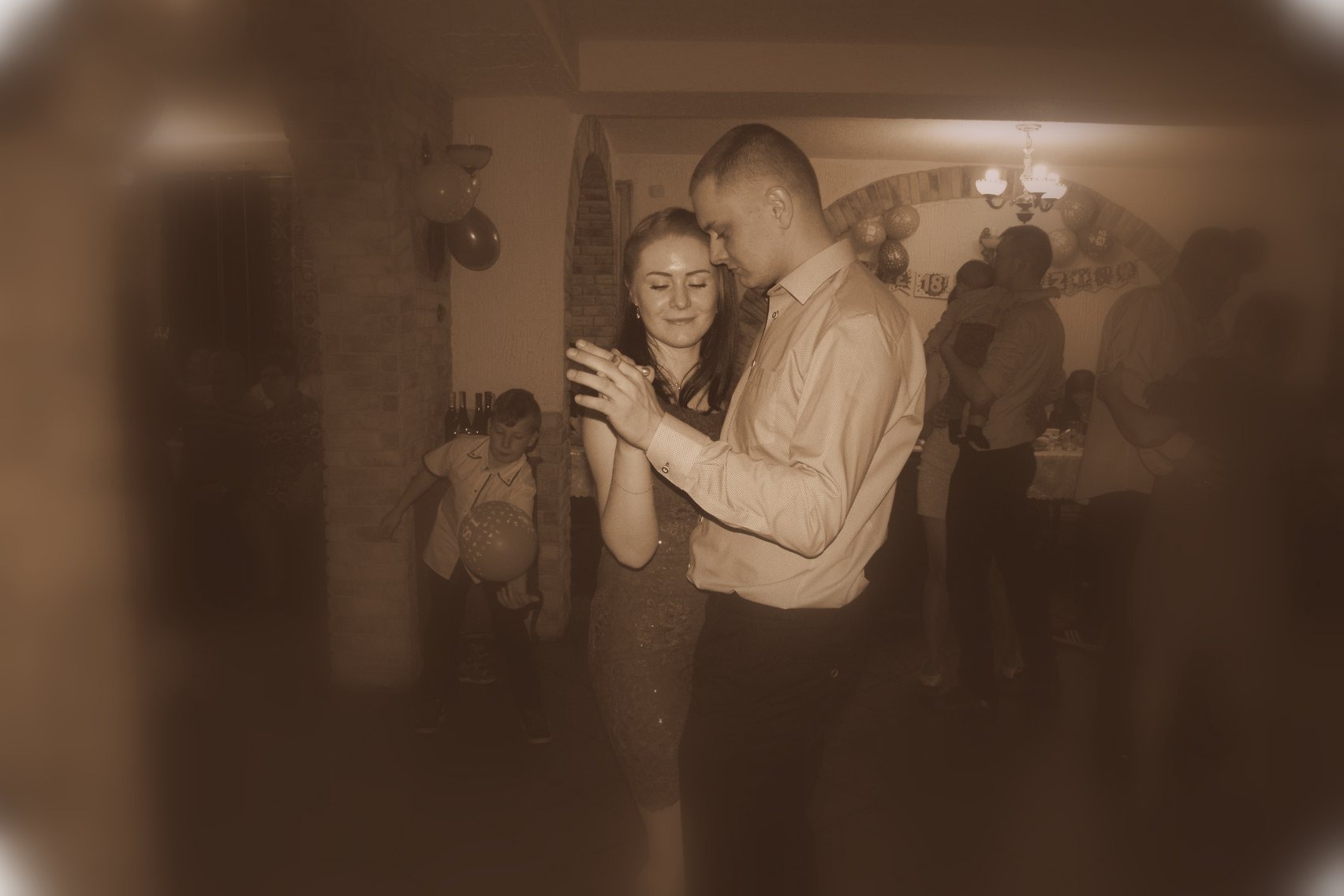 Para tańczy na tle sali udekorowanej balonami, w tle widoczni inni goście i dziecko z balonem.