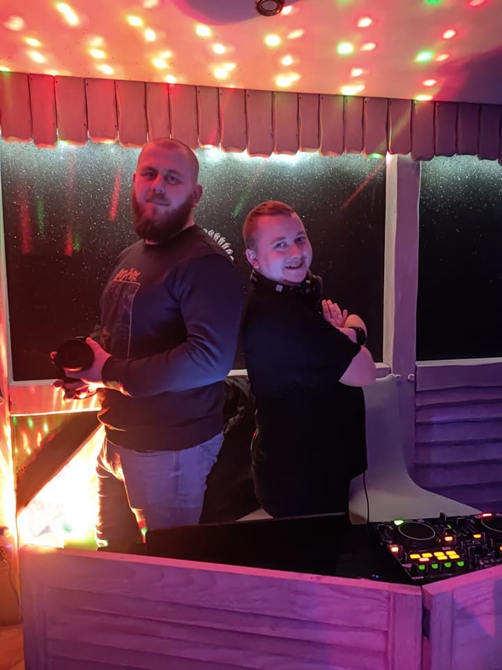 Dwóch mężczyzn za didżejką, jeden trzyma słuchawki, drugi z założonymi słuchawkami, oświetlenie dyskotekowe.