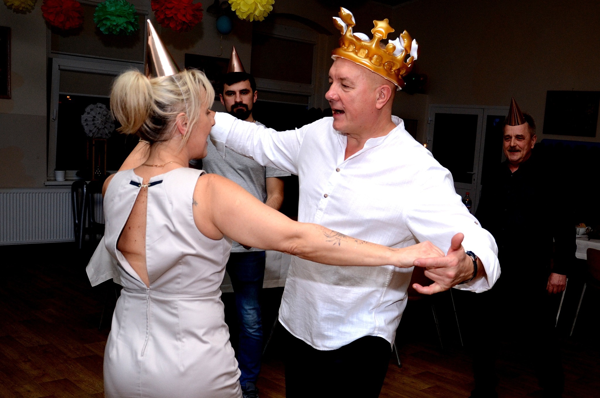 Para tańczy na imprezie, mężczyzna w koronie, kobieta w sukience z odkrytymi plecami, w tle osoby w czapkach urodzinowych i dekoracje.