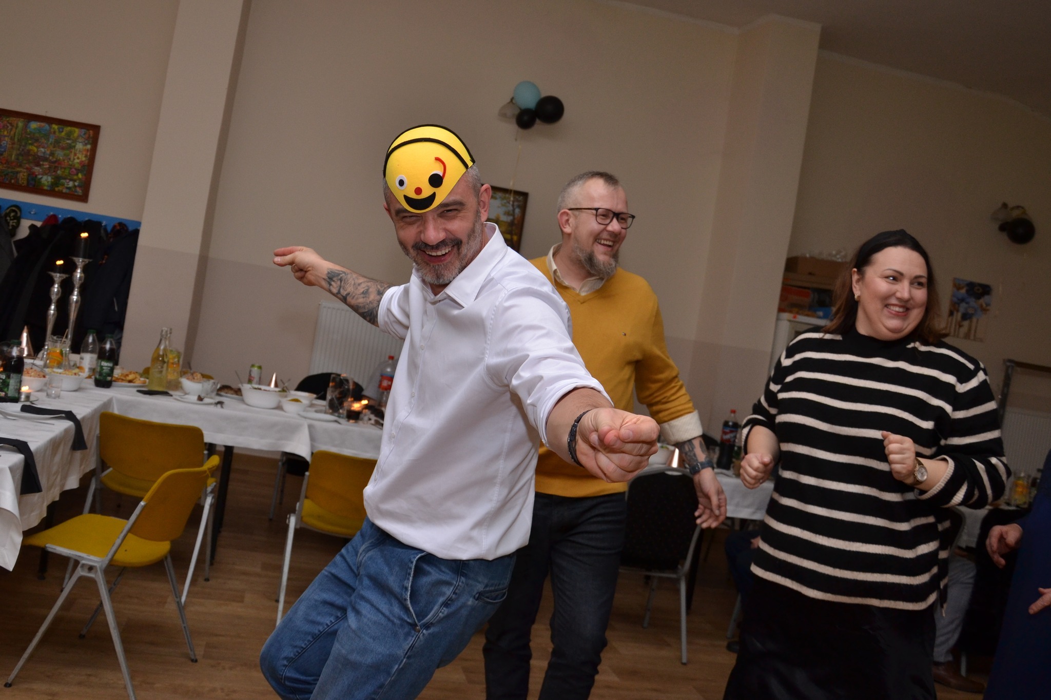 Uczestnicy imprezy tanecznej wesoło bawią się na parkiecie; mężczyzna w koszuli i jeansach ma na głowie nakrycie w kształcie uśmiechniętej pszczoły.