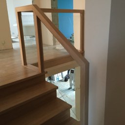 Greg-Mart Schody i Podłogi drewniane - Jasne, dębowe stopnie schodów z balustradą z drewna i szkła, widoczne w trakcie montażu, z pracownikiem w kombinezonie w tle.