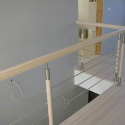 Greg-Mart Schody i Podłogi drewniane - Jasna balustrada z drewnianymi słupkami i metalowymi linkami na tle szarej ściany i podłogi z paneli, widoczny wentylator i wejście do łazienki.