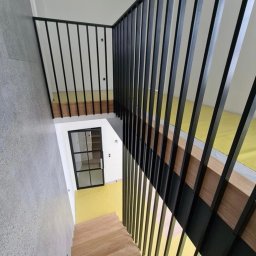 Greg-Mart Schody i Podłogi drewniane - Wysokie, proste schody z jasnego drewna z czarną, minimalistyczną balustradą z pionowych prętów, widok z góry na klatkę schodową z żółtą podłogą i przeszklonymi drzwiami na dole.