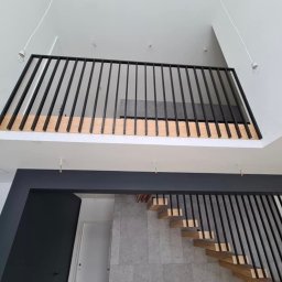Greg-Mart Schody i Podłogi drewniane - Nowoczesne wnętrze z drewnianymi schodami i czarną metalową balustradą, widok z dołu, szare płytki na ścianie, minimalizm.