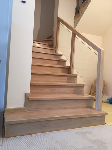 Jasne, dębowe schody z przeszkloną balustradą, widok z dołu na kilka stopni z widocznymi niedoskonałościami wykończenia podłogi.