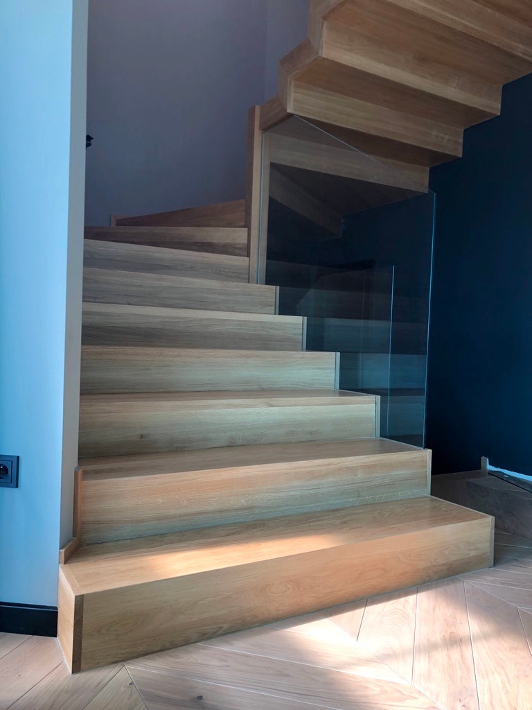 Nowoczesne, drewniane schody z przezroczystą balustradą szklaną, oświetlone naturalnym światłem, z parkietem ułożonym w jodełkę na podłodze.