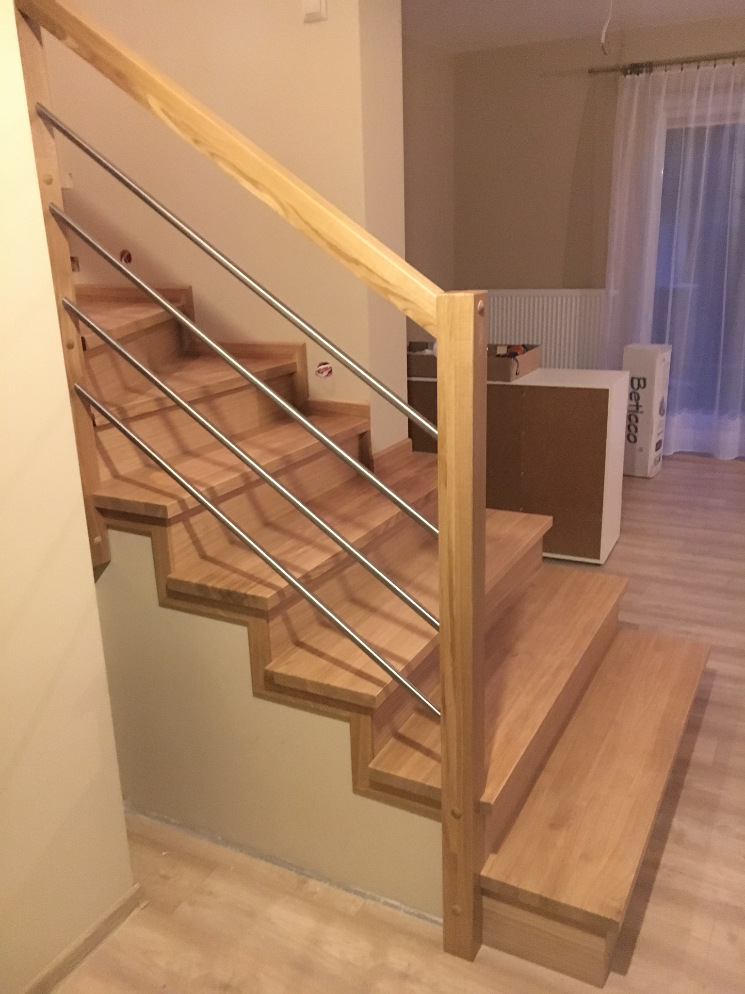 Jasne, drewniane stopnie schodów z metalową balustradą w nowoczesnym wnętrzu, widok z dołu na pierwsze stopnie, z elementami wykończeniowymi w tle.