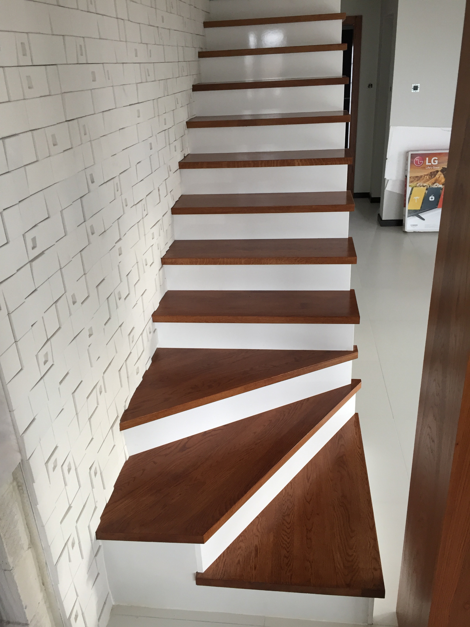 Bieg schodów z drewnianymi stopniami i białymi podstopnicami, widok z dołu, obok ściana z dekoracyjnymi panelami 3D, w tle widoczny fragment pomieszczenia.