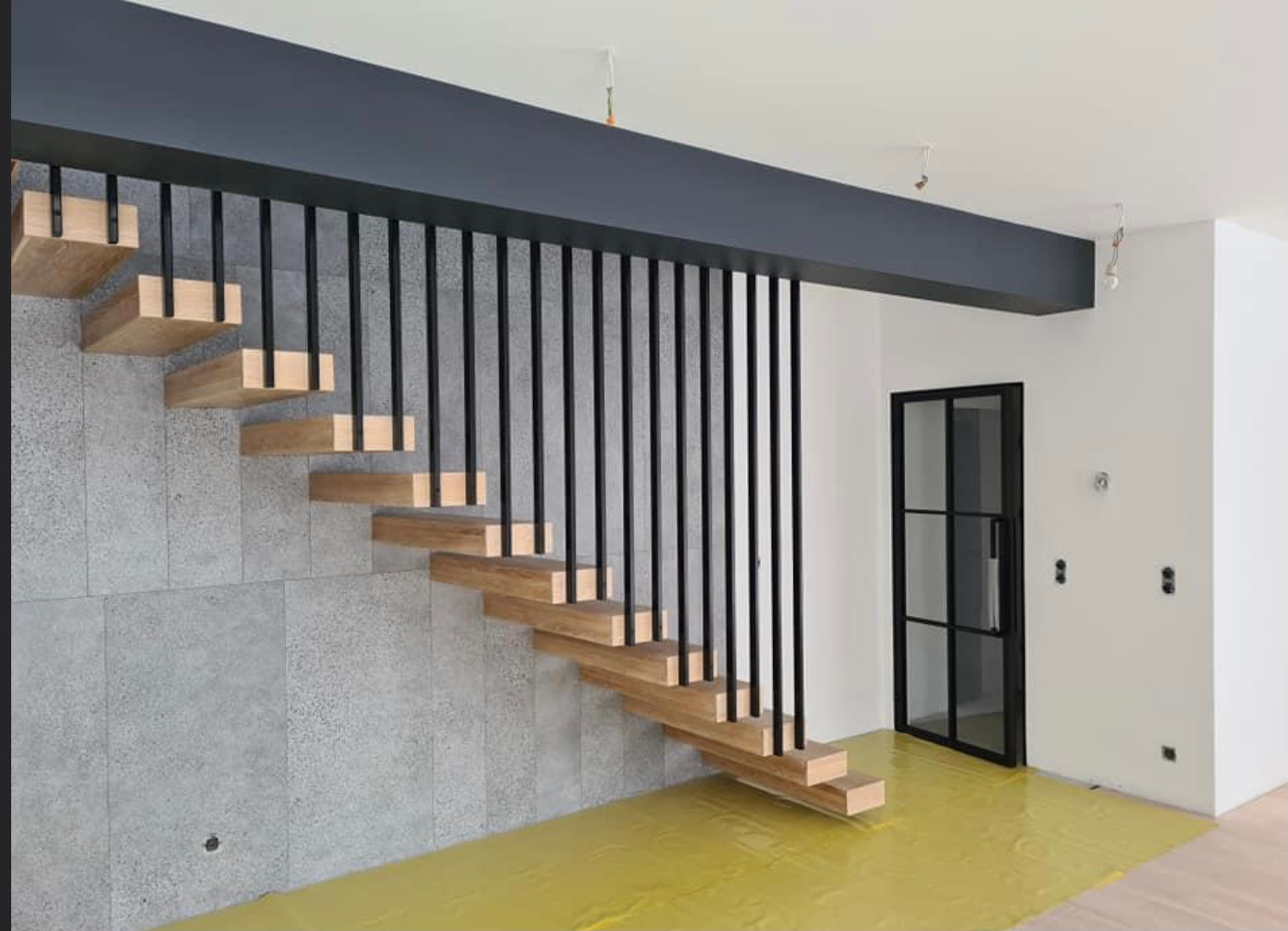 Nowoczesne schody wspornikowe z drewnianymi stopniami i czarną metalową balustradą, widoczne na tle szarej ściany i zabezpieczonej folią podłogi w trakcie montażu.