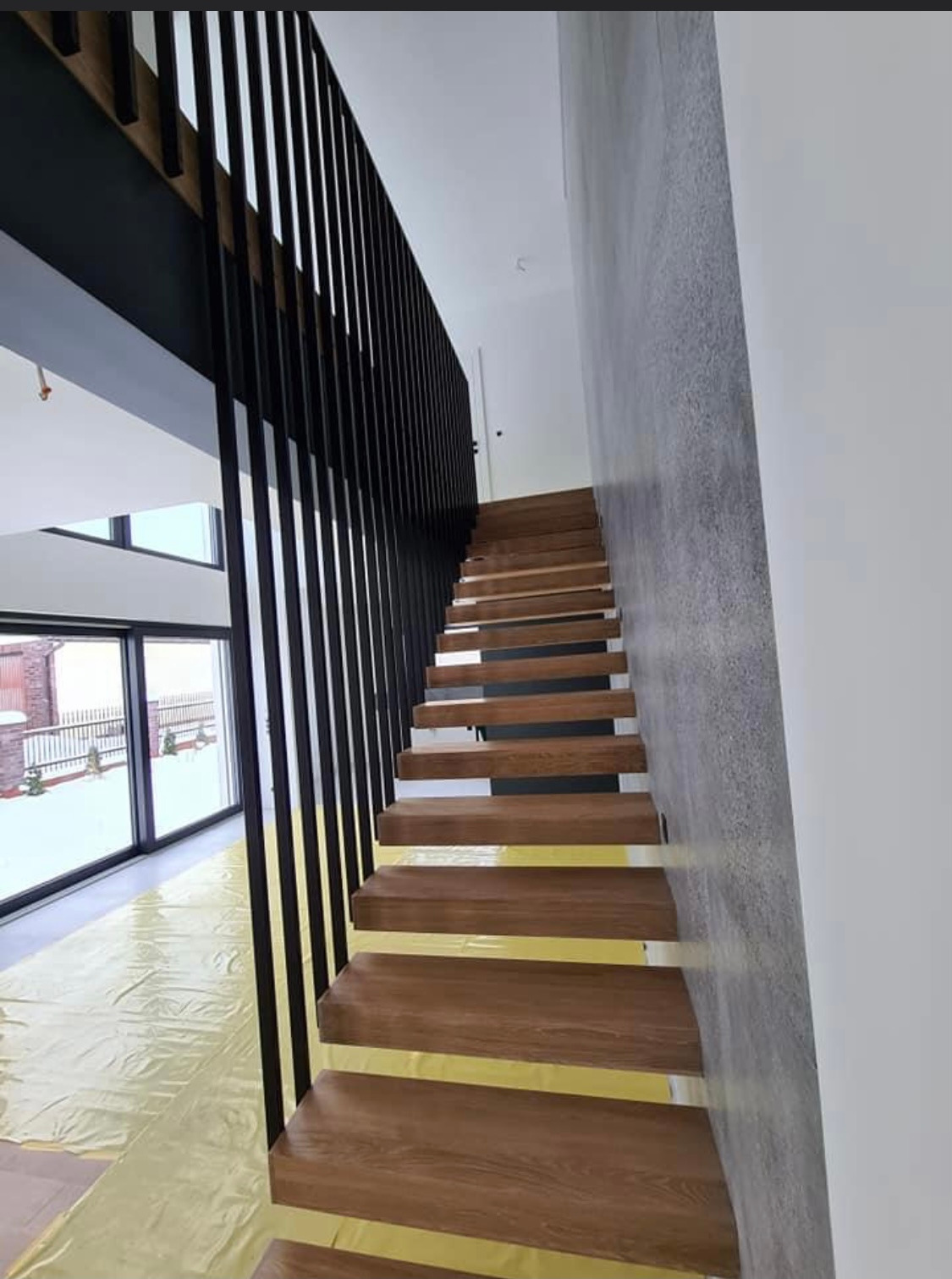 Nowoczesne schody drewniane z czarną metalową balustradą i szarą ścianą w tle, zabezpieczone folią ochronną.