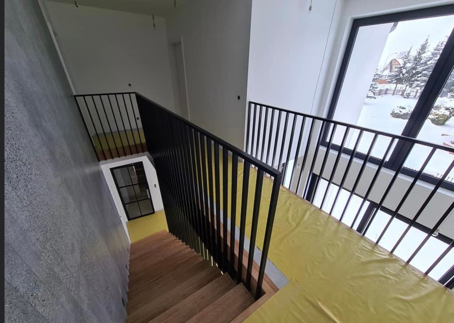 Wnętrze domu w trakcie wykańczania z widocznymi drewnianymi schodami z czarną metalową balustradą, zabezpieczone folią podłogi i okno z widokiem na zimowy krajobraz.