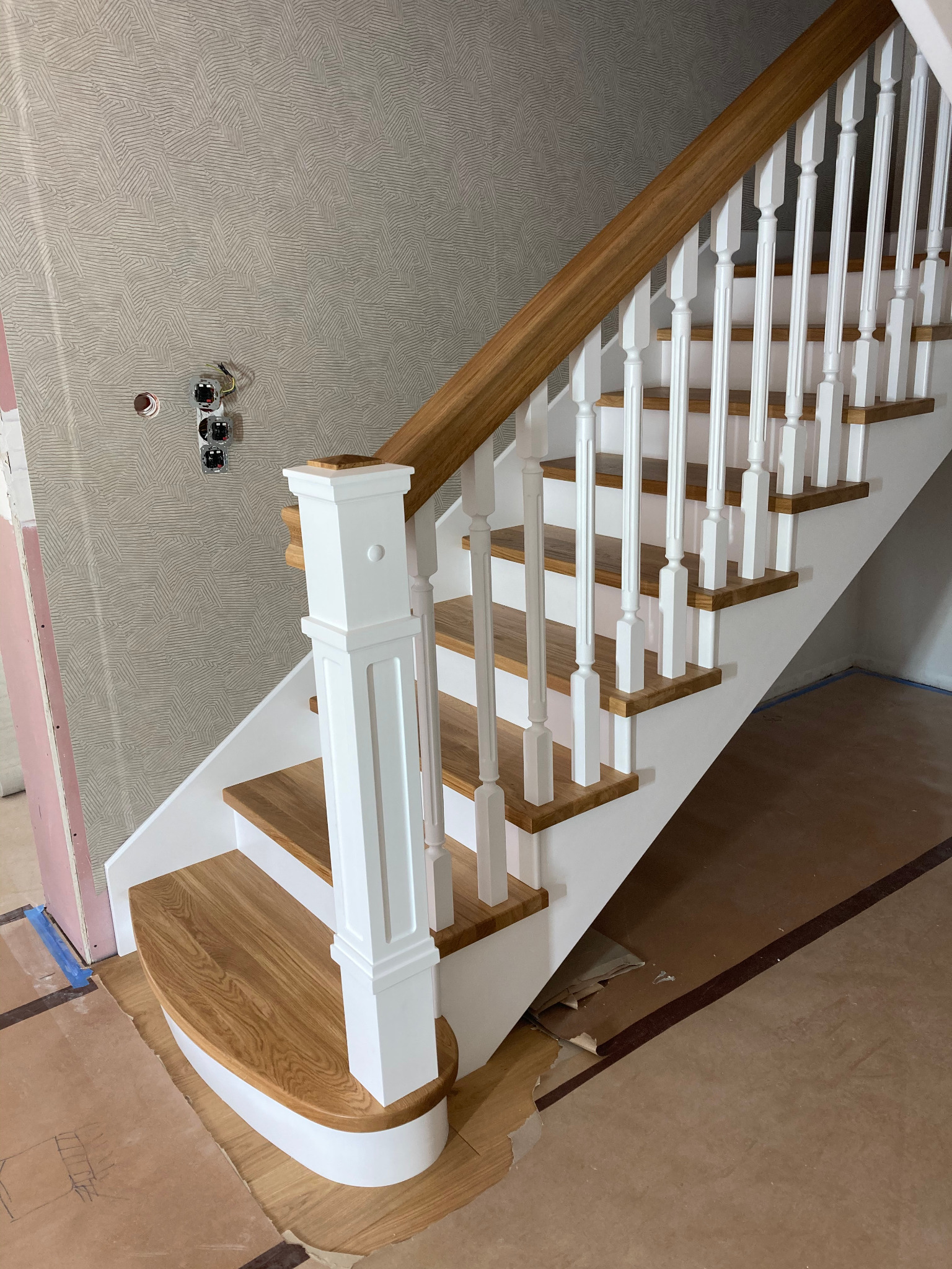Nowe schody z dębowymi stopniami i białą balustradą w trakcie montażu, widoczne niezabezpieczone kable elektryczne i karton na podłodze.