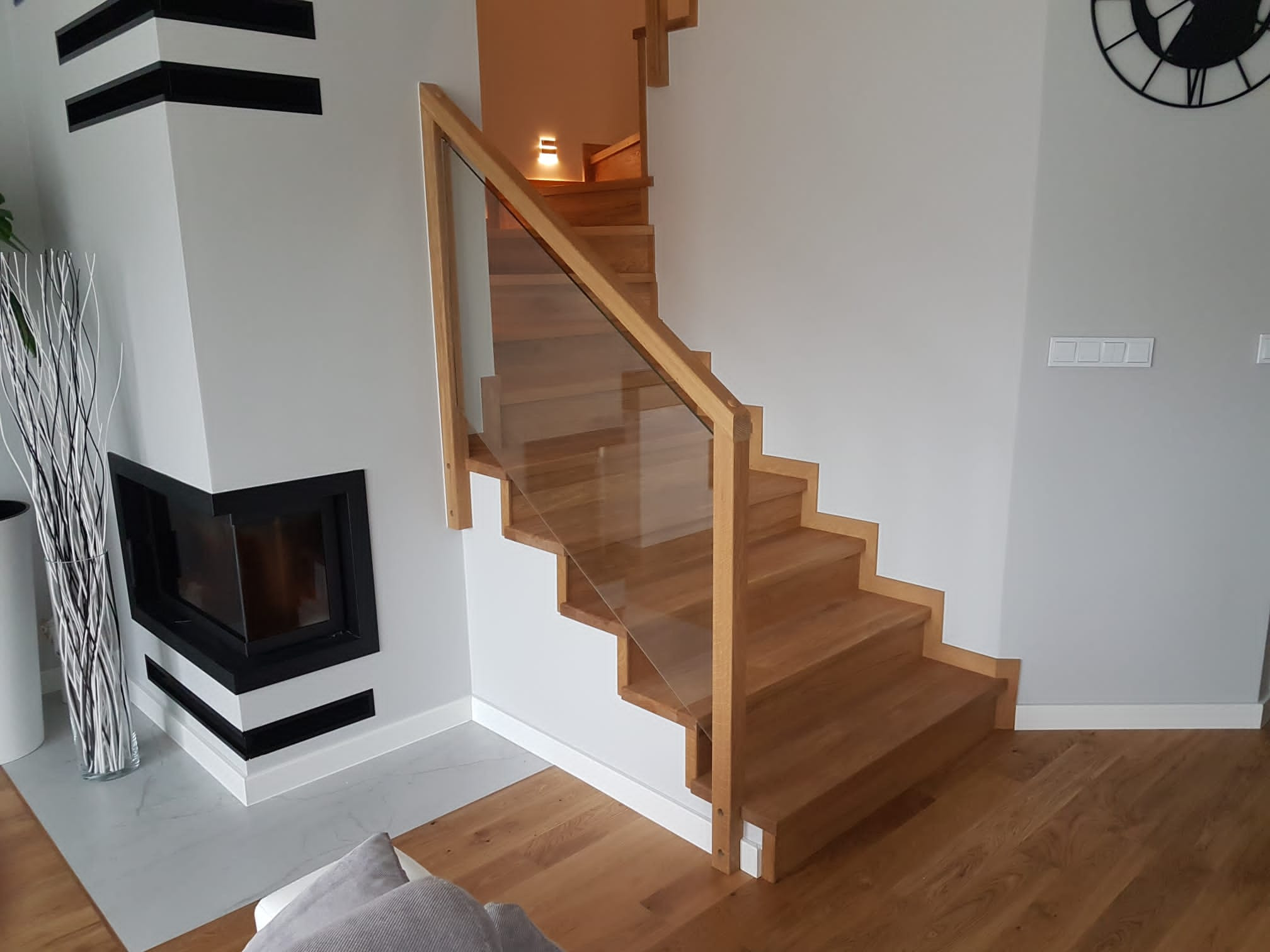 Jasne, drewniane schody z przezroczystą szklaną balustradą i drewnianą poręczą, w nowoczesnym wnętrzu z kominkiem i zegarem na ścianie.
