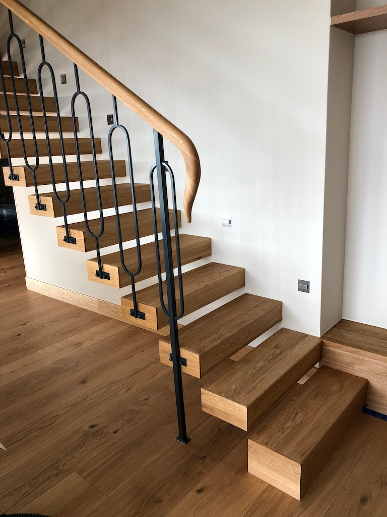 Nowoczesne schody z drewnianymi stopniami i czarną, metalową balustradą o minimalistycznym designie, połączone z drewnianą podłogą o ciepłym odcieniu, oświetlone naturalnym światłem.