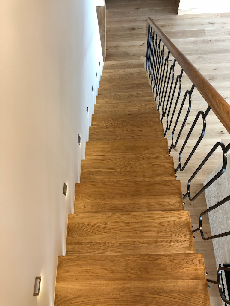 Dębowe schody z czarną metalową balustradą o dekoracyjnych zaokrągleniach, oświetlone małymi lampami ściennymi, widok z góry.