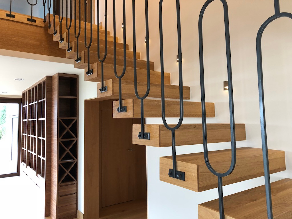 Nowoczesne, dębowe schody wspornikowe z czarną, metalową balustradą o minimalistycznym kształcie, podświetlone punktowymi lampkami ściennymi, z widocznym regałem na wino w tle.