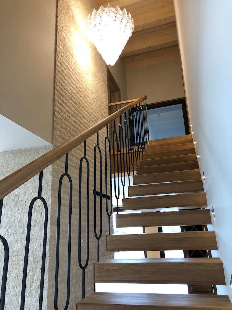 Nowoczesne schody z drewnianymi stopniami i czarną, metalową balustradą o minimalistycznym wzorze, oświetlone małymi punktowymi lampkami, prowadzące do góry w jasnym wnętrzu z kamienną ścianą...