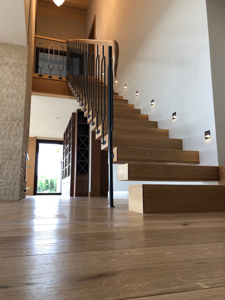 Nowoczesne schody z drewnianymi stopniami i czarną metalową balustradą, oświetlone punktowymi lampami na ścianie, widok z poziomu podłogi.