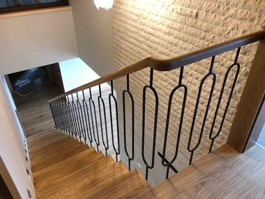 Widok z góry na dębowe schody z czarną, metalową balustradą o owalnych prętach, z jasną ścianą wyłożoną kamieniem dekoracyjnym w tle.
