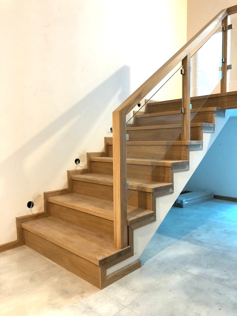 Jasne, dębowe schody z szklaną balustradą, widoczne otwory na puszki elektryczne w ścianie, surowy stan wykończenia.