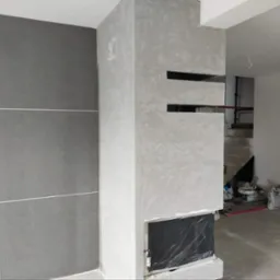 Ściana z dekoracyjną szarą strukturą, imitująca kamień, z ciemniejszymi płytami oddzielonymi białymi fugami po lewej stronie, fragment kominka w trakcie wykończenia z widocznymi otworami, surowy...