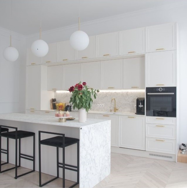 Jasna kuchnia z białymi, frezowanymi frontami, złotymi uchwytami i marmurowym blatem wyspy, oświetlona kulistymi lampami wiszącymi, z bukietem czerwonych piwonii na wyspie.