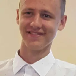 Portret uśmiechniętego młodego mężczyzny w białej koszuli w kropki, z krótkimi włosami i niewielką blizną na czole.