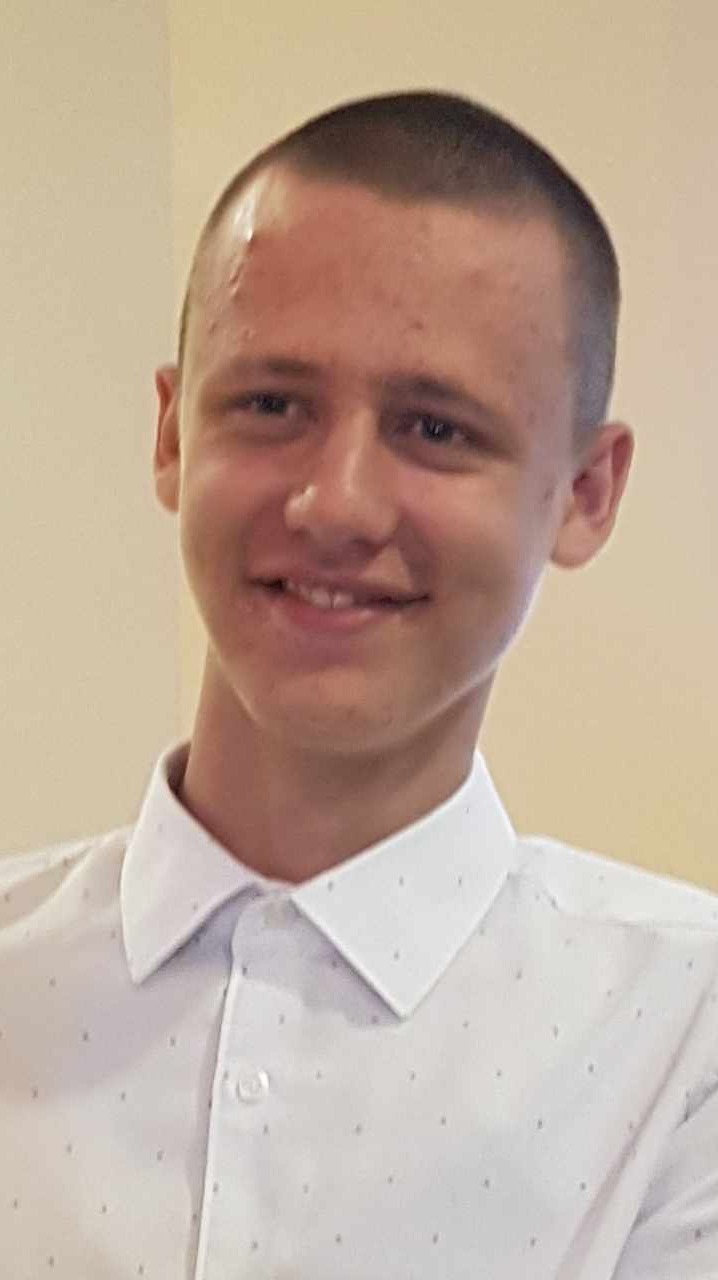 Portret uśmiechniętego młodego mężczyzny w białej koszuli w kropki, z krótkimi włosami i niewielką blizną na czole.