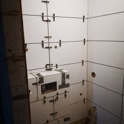 Andrzej Pietrzkiewicz - Ściany łazienki w trakcie układania białych, prostokątnych płytek z wykorzystaniem krzyżyków dystansowych, widoczny fragment surowej ściany i instalacja podtynkowa zabezpieczona taśmą.