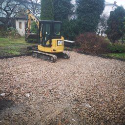 Andrzej Pietrzkiewicz - Żółta mini koparka CAT na placu budowy, przygotowanym podłoże z gruzu, widok na ogród z budynkami w tle.