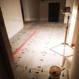 Andrzej Pietrzkiewicz - Szare płytki podłogowe w trakcie układania, widoczne krzyżyki dystansowe, poziomica, wiadro i lampa halogenowa na trójnogu, niedokończone ściany w tle.