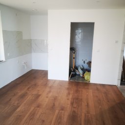 Andrzej Pietrzkiewicz - Brązowe panele podłogowe ułożone w jasnym pomieszczeniu z fragmentami wykończonej ściany z marmurowymi płytkami i przygotowanym otworem na drzwi, w którym widoczne są narzędzia i materiały budowlane.