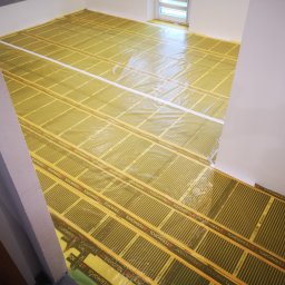 Andrzej Pietrzkiewicz - Montaż folii grzewczej Termofol na podłodze w pomieszczeniu z białymi ścianami, przygotowanie do układania paneli podłogowych, widoczne taśmy klejące i fragment okna.