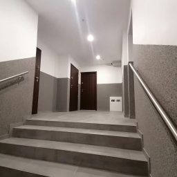 Andrzej Pietrzkiewicz - Klatka schodowa z szarymi płytkami na stopniach, metalowa poręcz po prawej stronie, trzy ciemnobrązowe drzwi w tle i biała szafka elektryczna.