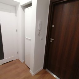 Andrzej Pietrzkiewicz - Wnętrze przedpokoju z dwiema parami drzwi: białe drzwi z czarnym paskiem oraz ciemnobrązowe drzwi z metalowymi okuciami, widoczny parkiet i domofon na ścianie.