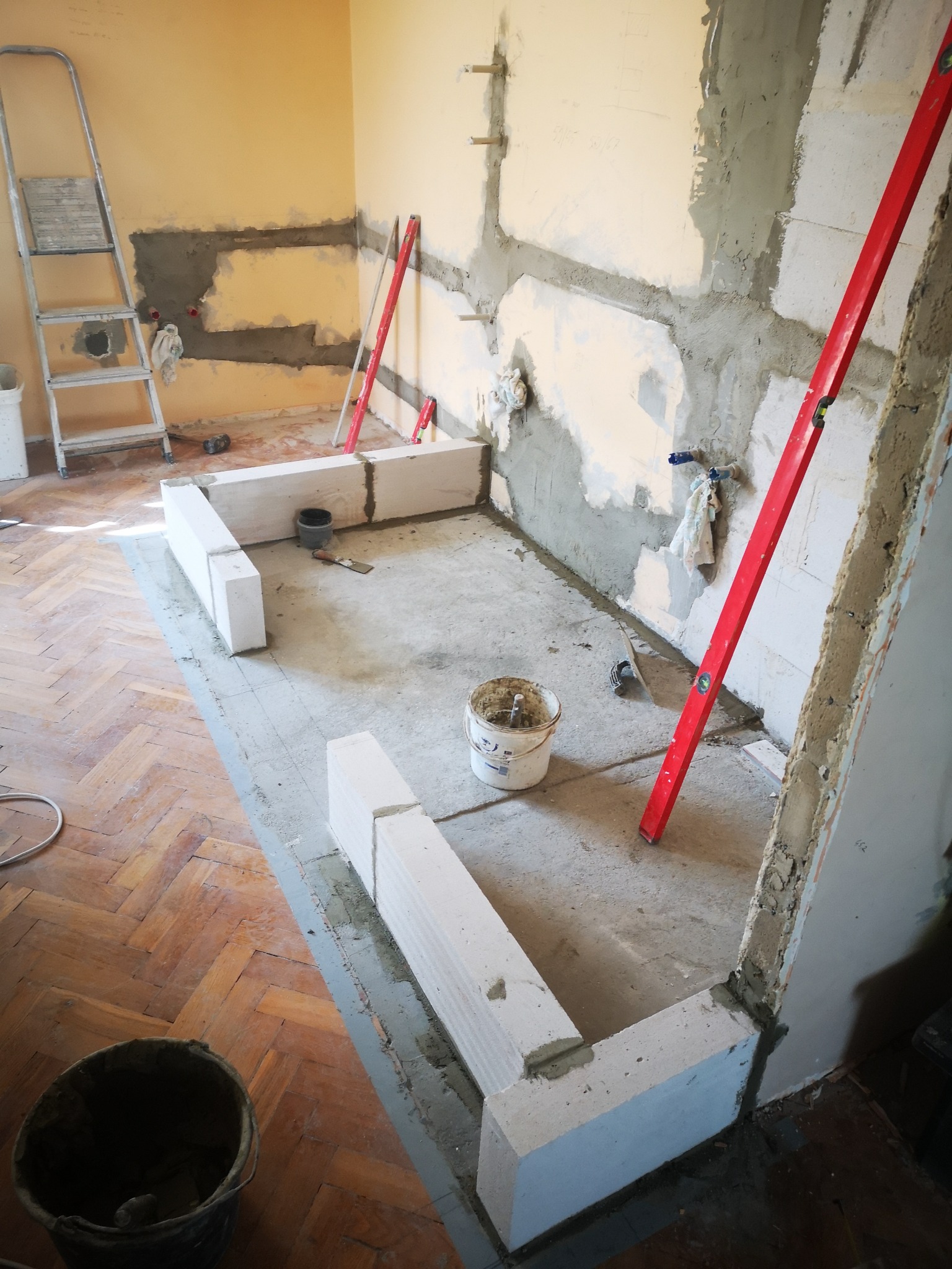 Wnętrze łazienki w trakcie remontu: widoczne bloczki z betonu komórkowego tworzące obrys brodzika, poziomica oparta o ścianę, ślady po skuciu tynku, drabina i wiadro z zaprawą.