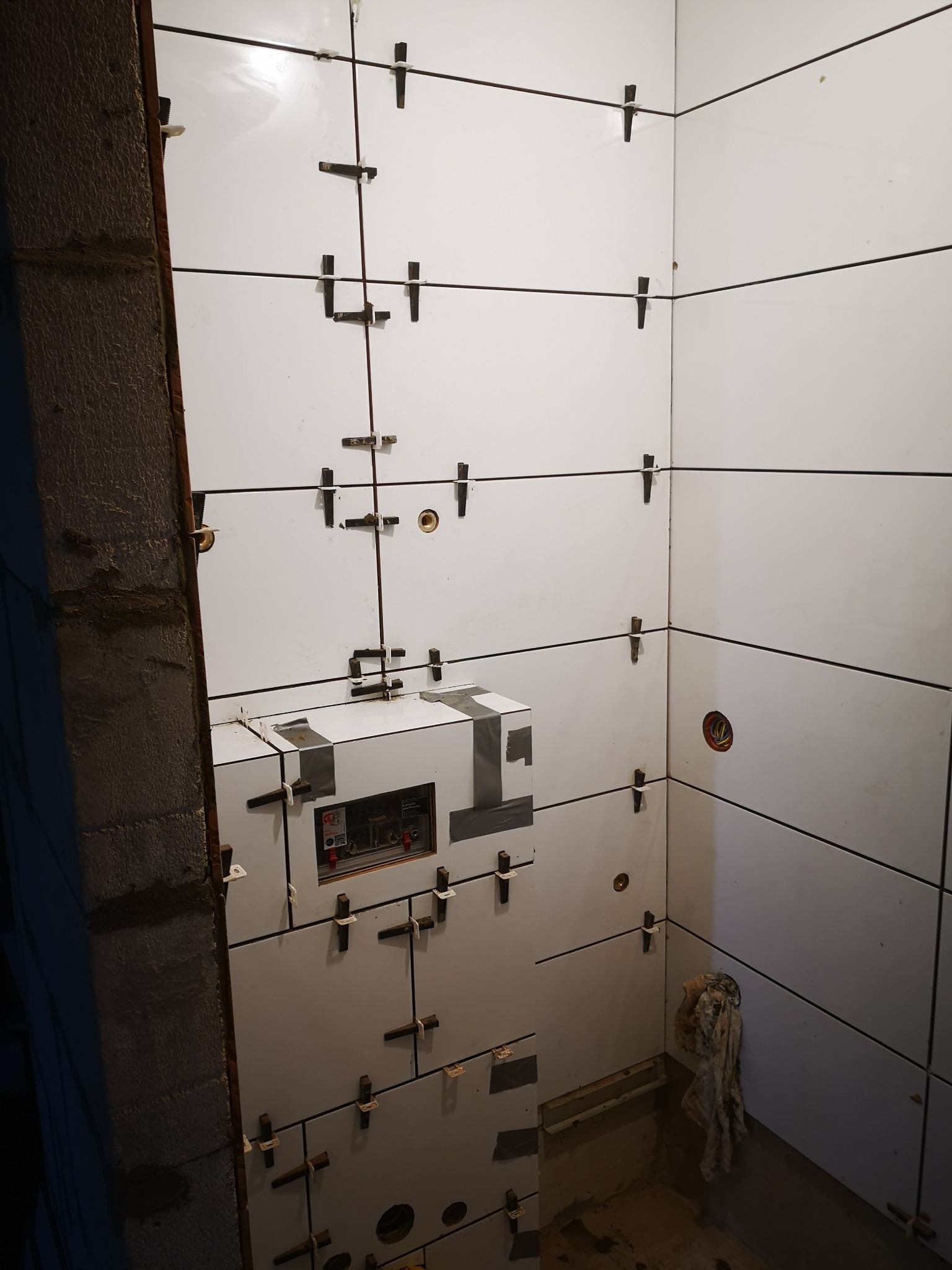 Ściany łazienki w trakcie układania białych, prostokątnych płytek z wykorzystaniem krzyżyków dystansowych, widoczny fragment surowej ściany i instalacja podtynkowa zabezpieczona taśmą.