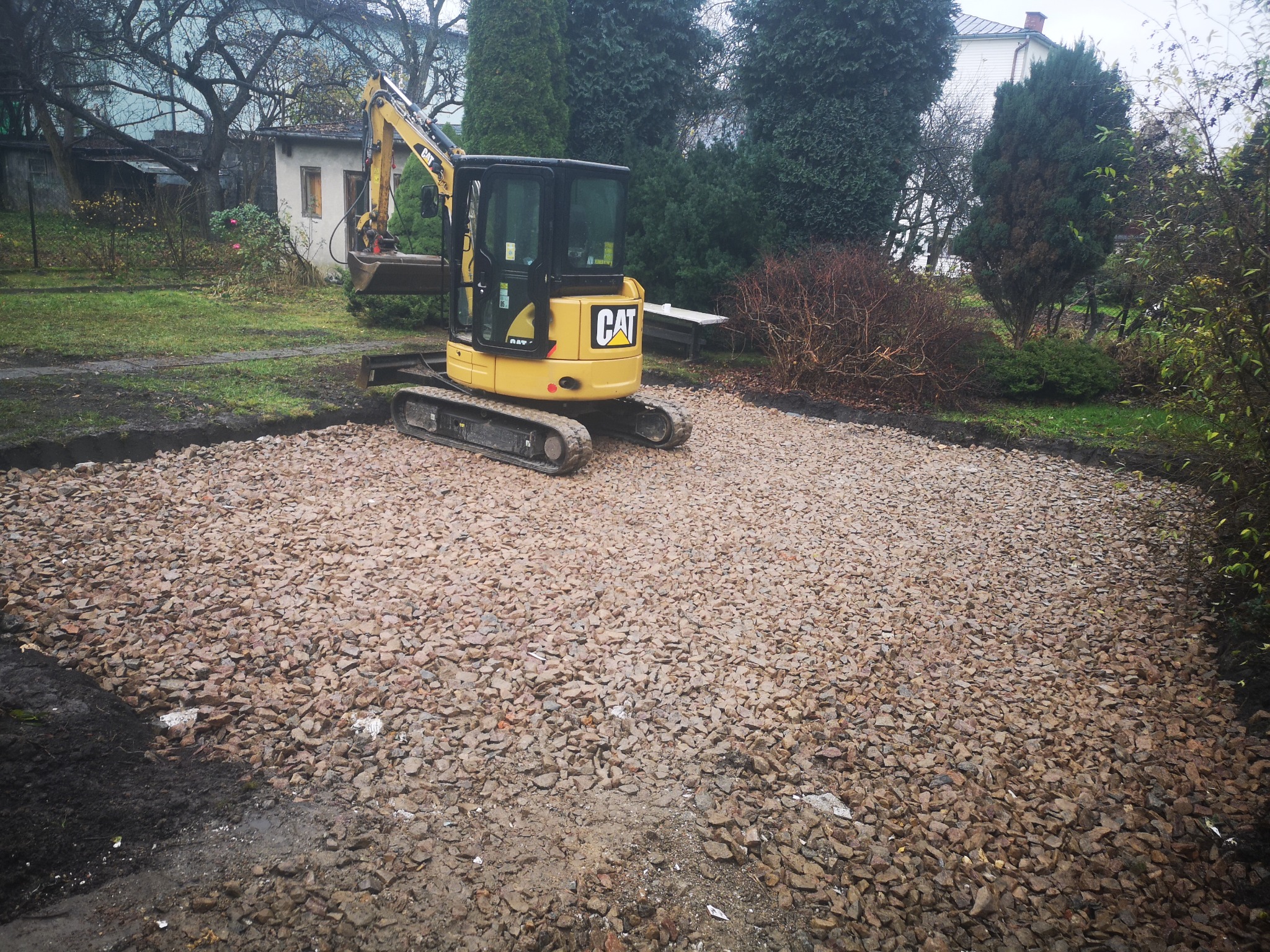 Żółta mini koparka CAT na placu budowy, przygotowanym podłoże z gruzu, widok na ogród z budynkami w tle.