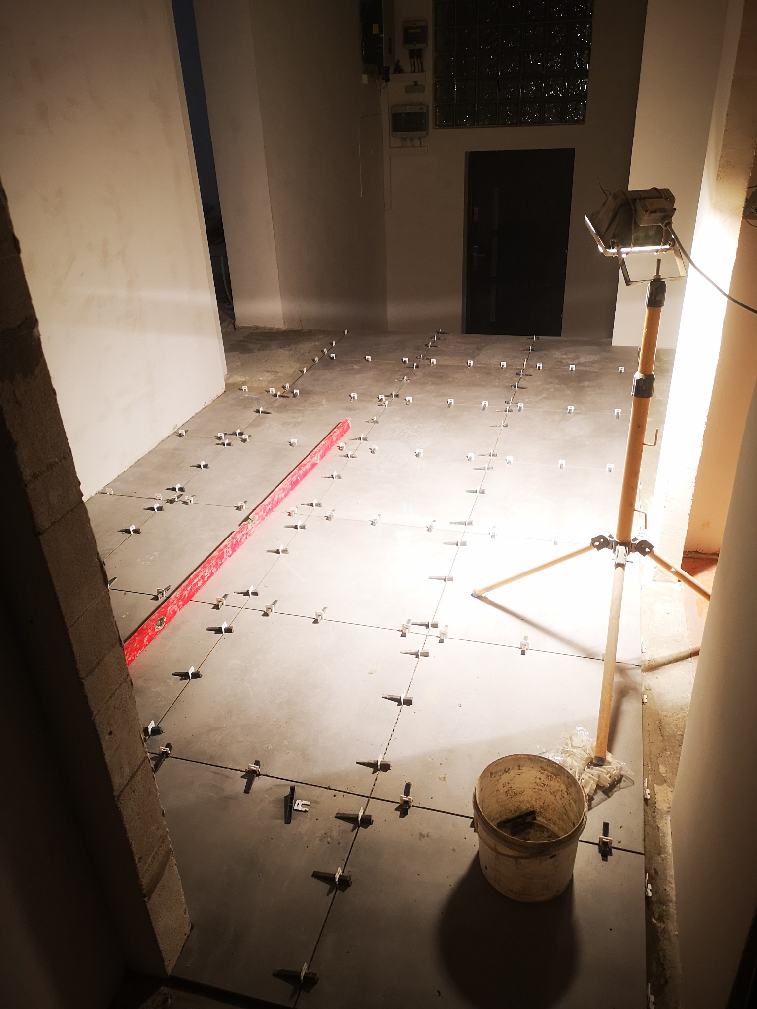 Szare płytki podłogowe w trakcie układania, widoczne krzyżyki dystansowe, poziomica, wiadro i lampa halogenowa na trójnogu, niedokończone ściany w tle.