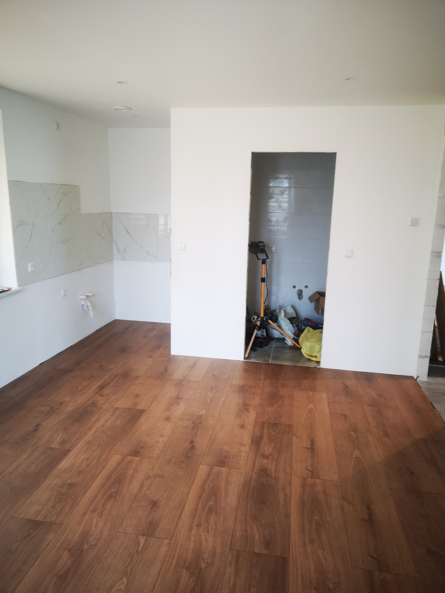 Brązowe panele podłogowe ułożone w jasnym pomieszczeniu z fragmentami wykończonej ściany z marmurowymi płytkami i przygotowanym otworem na drzwi, w którym widoczne są narzędzia i materiały budowlane.