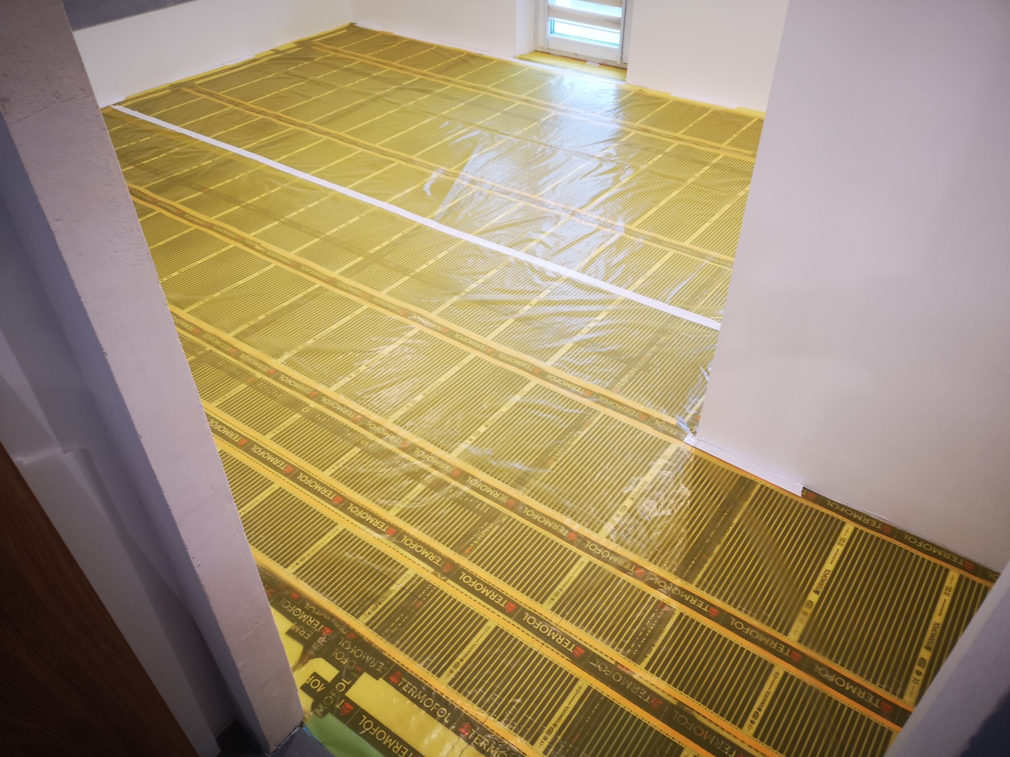 Montaż folii grzewczej Termofol na podłodze w pomieszczeniu z białymi ścianami, przygotowanie do układania paneli podłogowych, widoczne taśmy klejące i fragment okna.