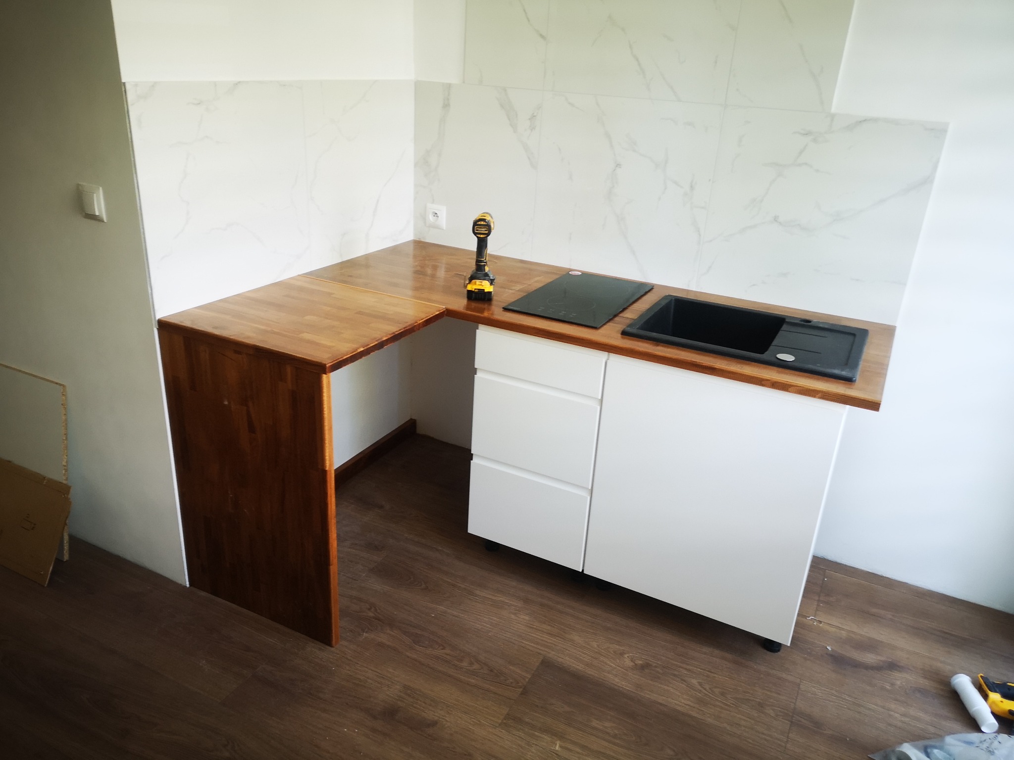 Kącik kuchenny z drewnianym blatem i białymi szafkami, czarny zlew i płyta indukcyjna, wiertarka akumulatorowa leży na blacie, białe płytki imitujące marmur na ścianie.