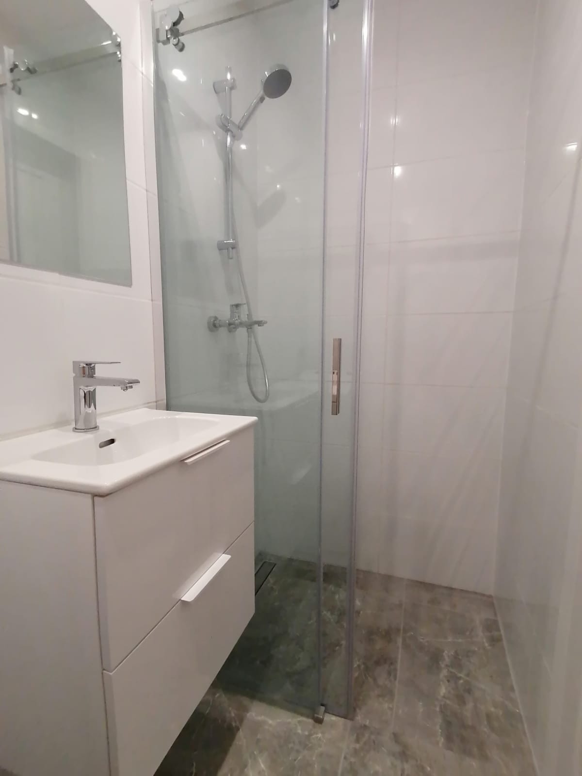 Nowoczesna łazienka w Wieliczce z białą szafką pod umywalkę z chromowaną baterią i kabiną prysznicową ze szklanymi drzwiami, wykończona białymi płytkami ściennymi i podłogą imitującą marmur.